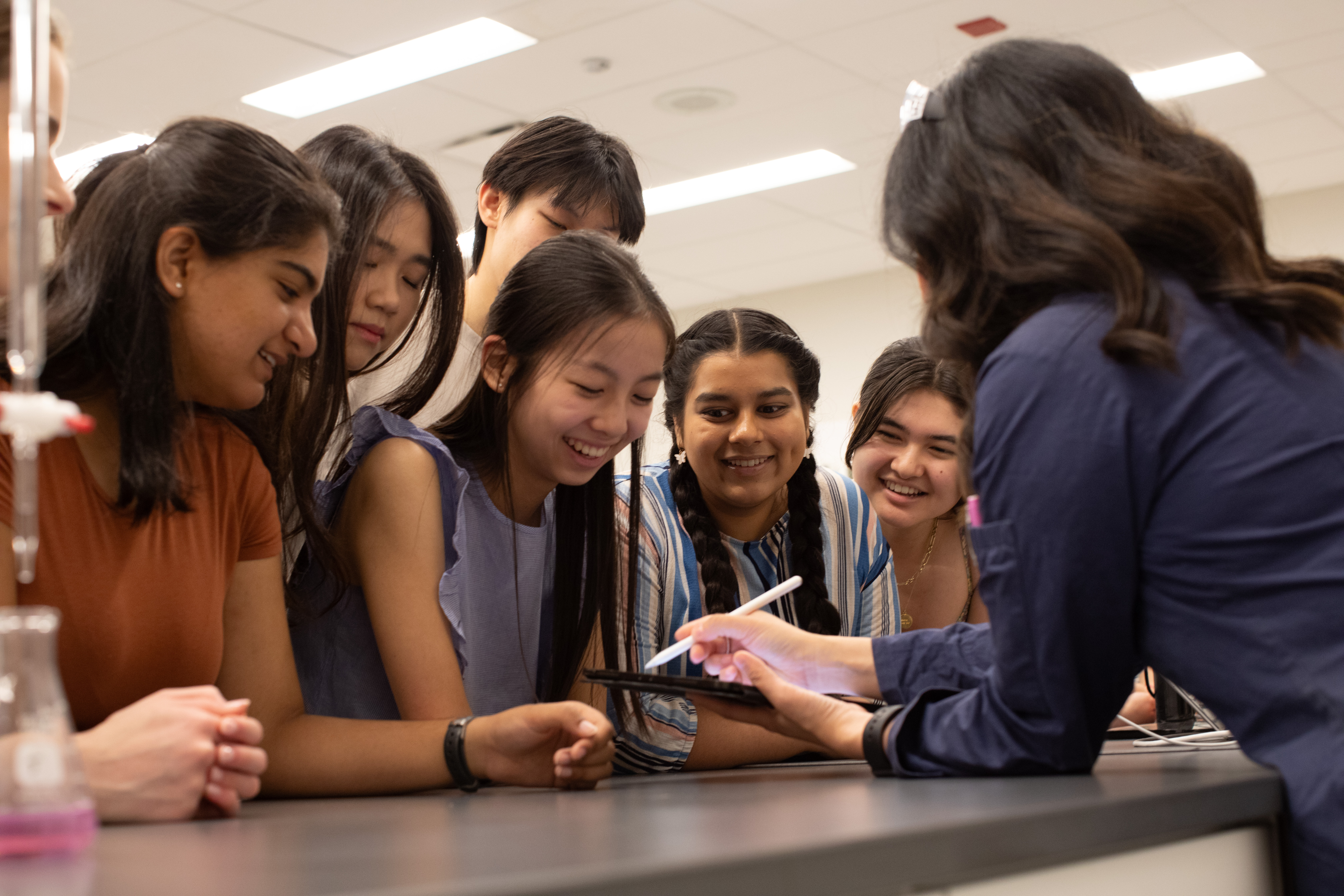 Students stand around a teacher who is teaching them a lesson on a tablet.