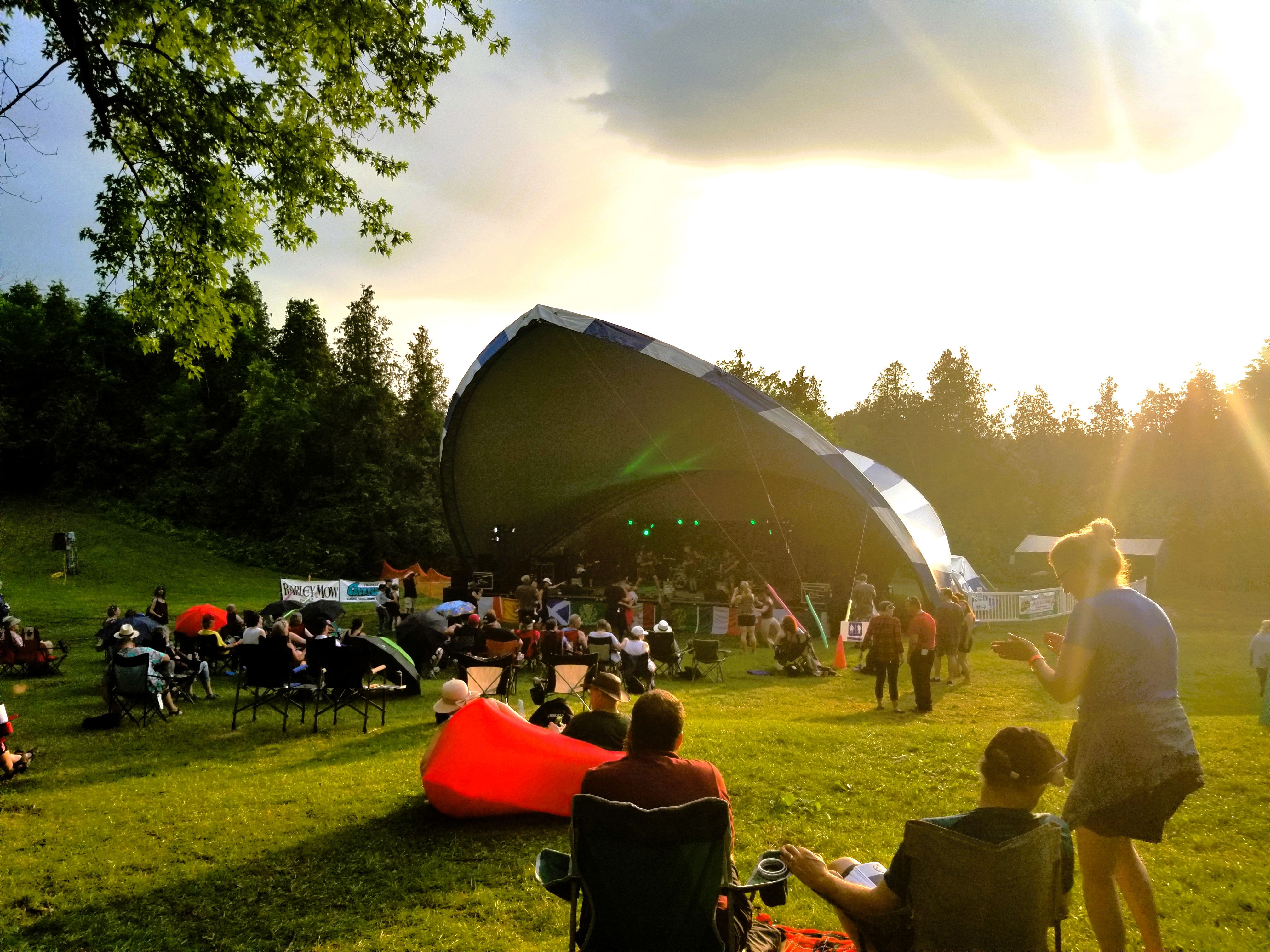 People sit in a grassy park area in lawn chairs near a bandshell, listening to performers on stage.