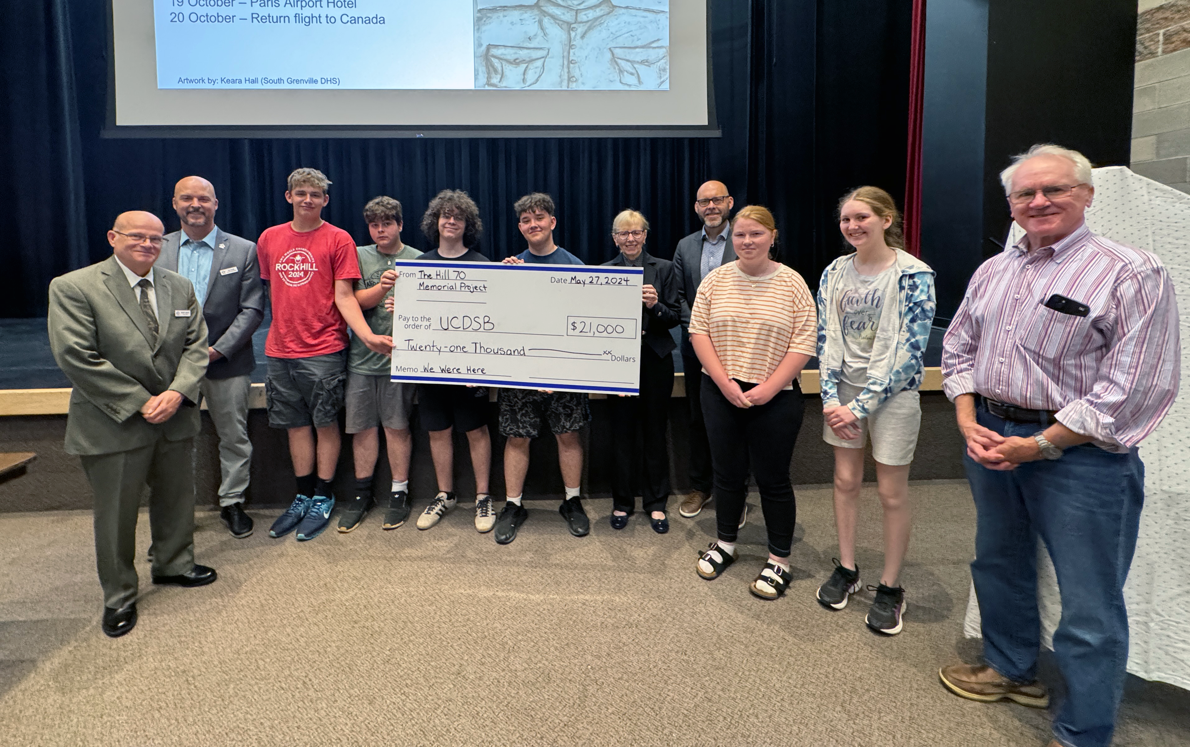 A group of people stand in a room together holding a large cheque, with a projector screen in the background