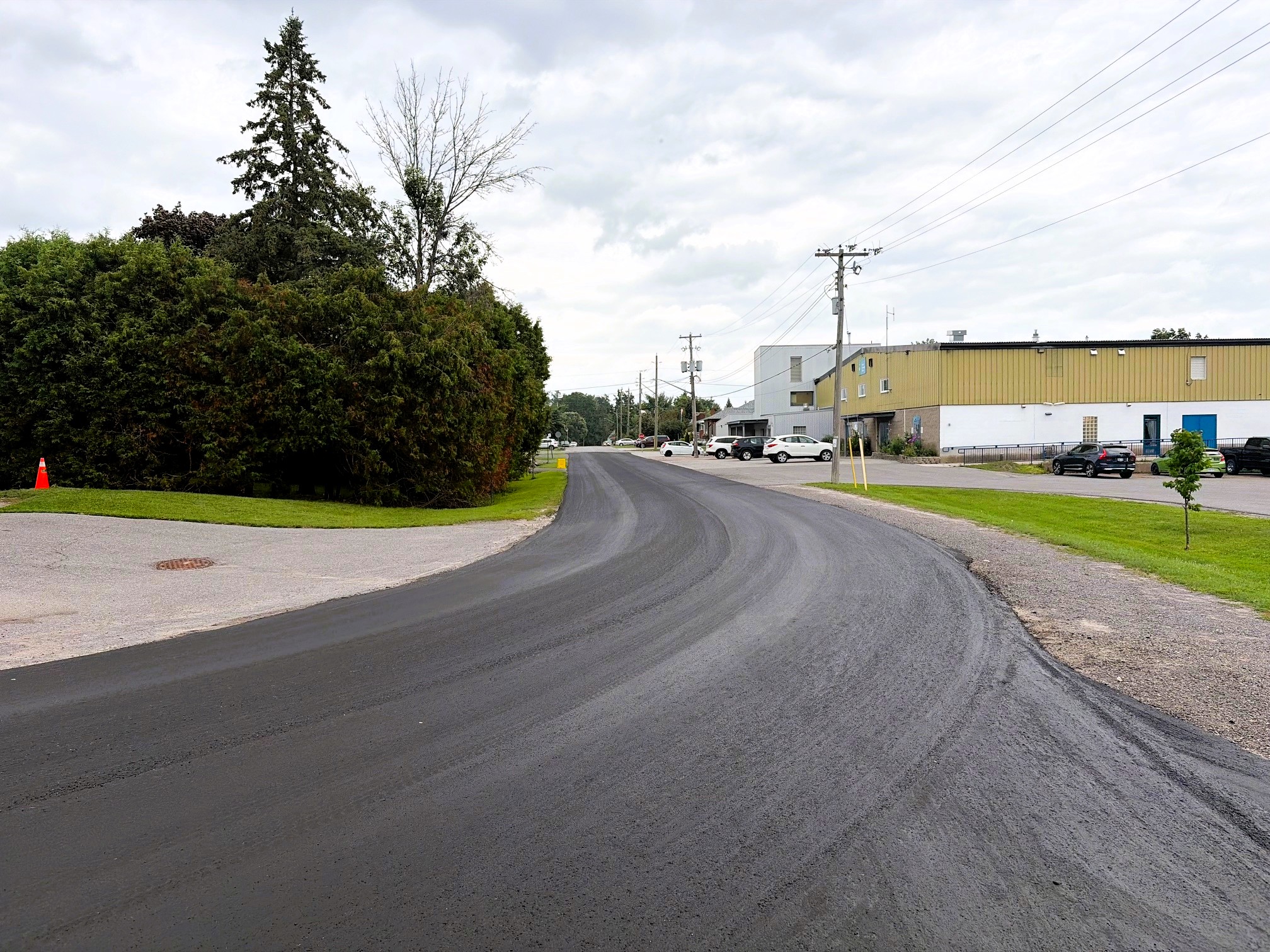 A freshly paved road in a village. An arena stands to the right, bushes to the left.