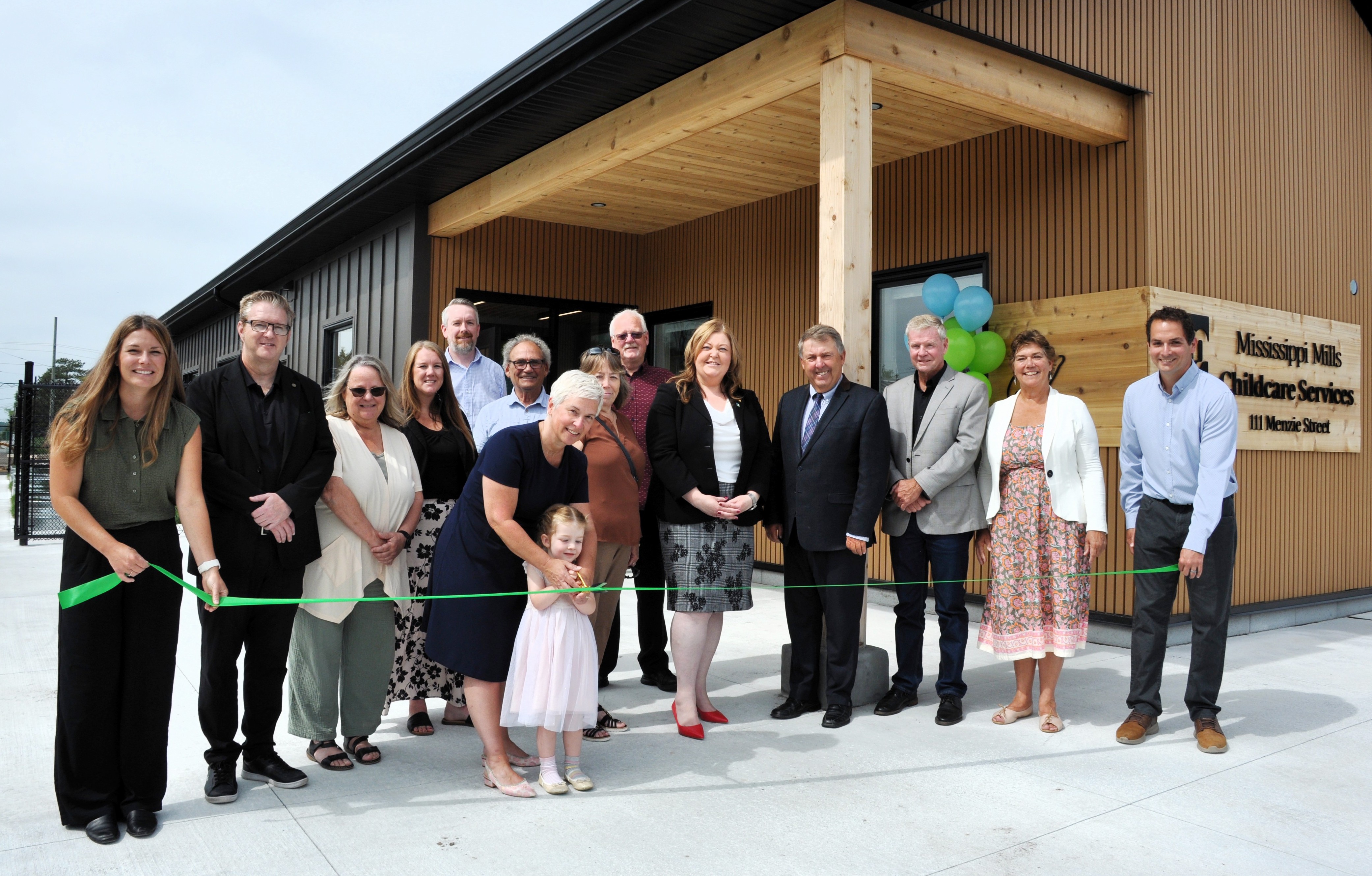 People stand together for a group photo in front of a new building. People in front hold a green ribbon on each end while a little prepares to cut the ribbon
