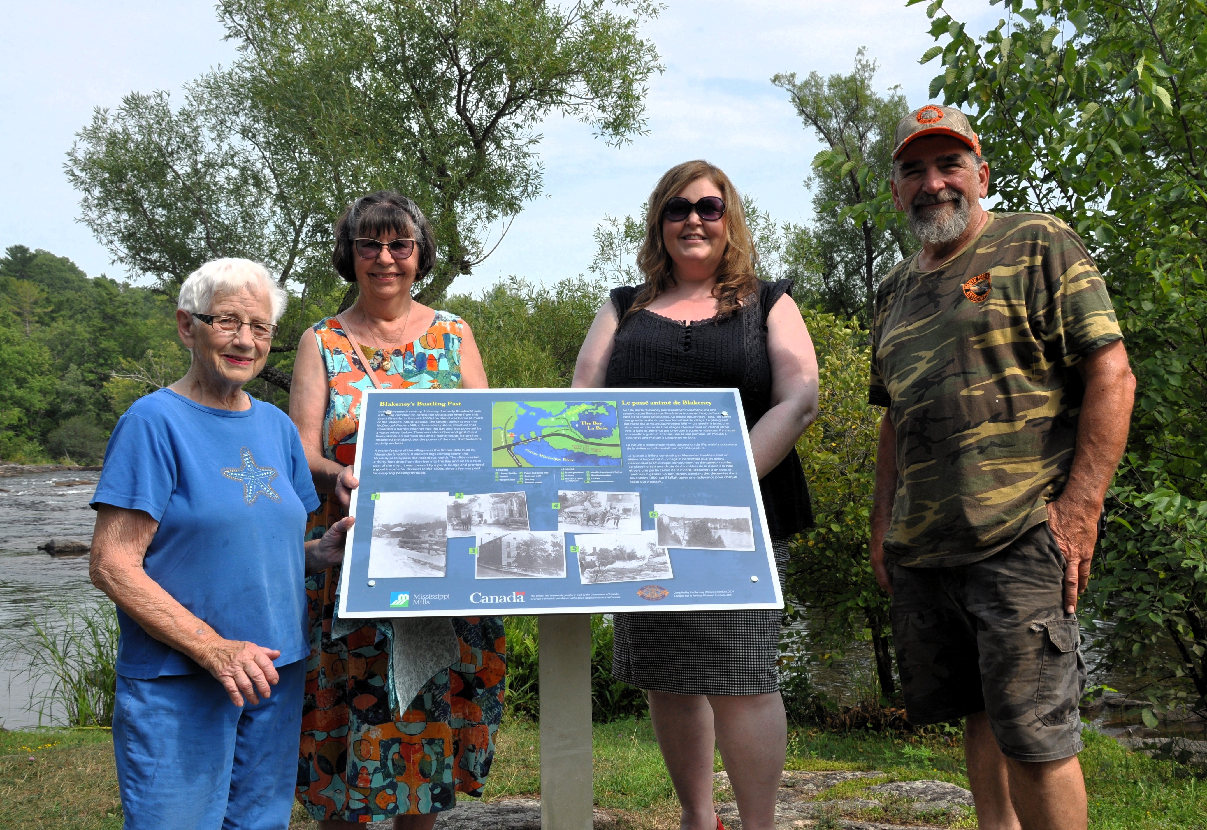 On a sunny day at the water's edge, four people gather around a blue plaque for a photo