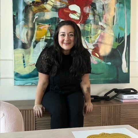Woman wearing black with long dark hair sits on the edge of a desk and smiles at the camera. A colourful painting is in the background.