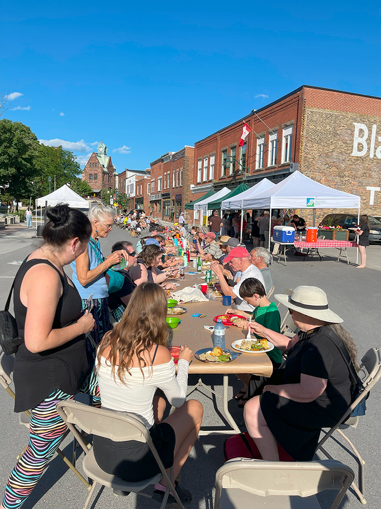 A group of people sit at tables lining a downtown street for a potluck.