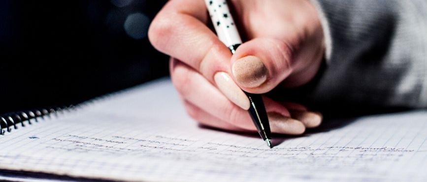 Hand holding a pen, writing in a spiral bound book.