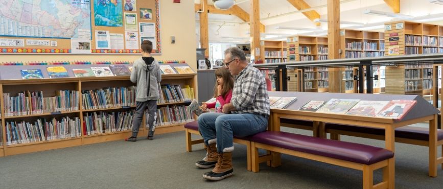 Young boy with his browsing a picture book, while a young girl and a man look at a book together, while seated on a small bench in the Port Carling branch.