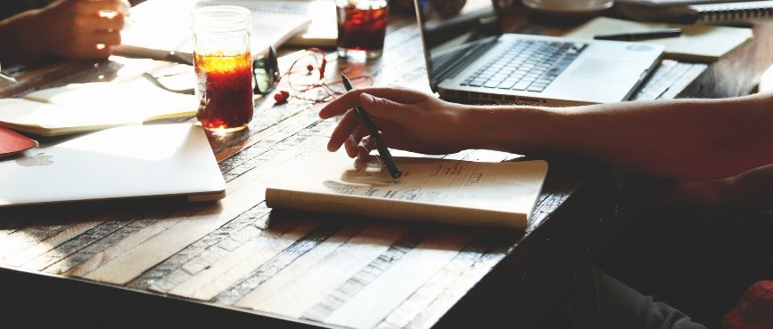 people using table for a meeting. One person is holding a pen, there are drinks on the table and a laptop.