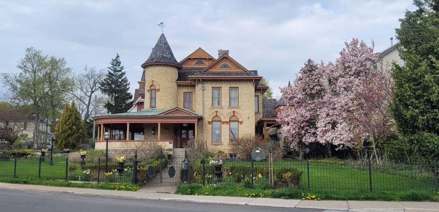 Beautiful old home with rounded covered porch and pointed peak, surrounded by green landscaping and a pink blossom tree.