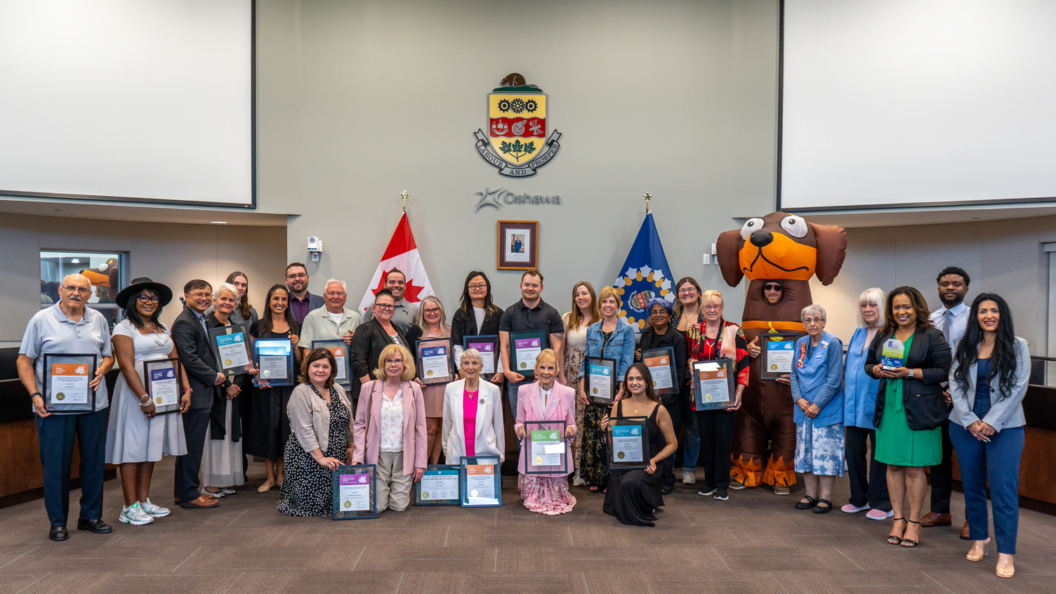 A large group of people stand together holding award certificates in City Council Chamber.