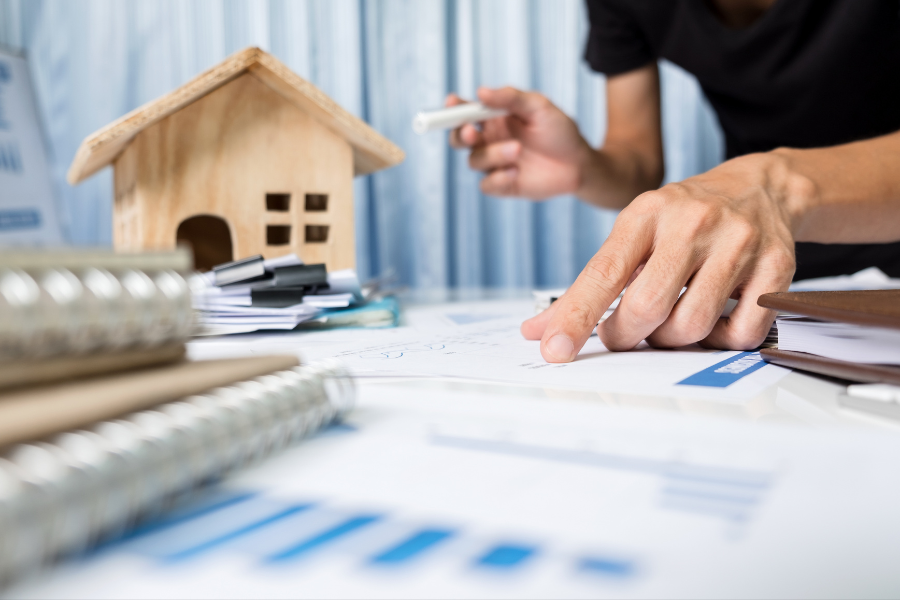 Small model of a house next to a person holding a pen in one hand and pointing at a document on the table with the other. Several documents are in two stacks on the table in front of the house.