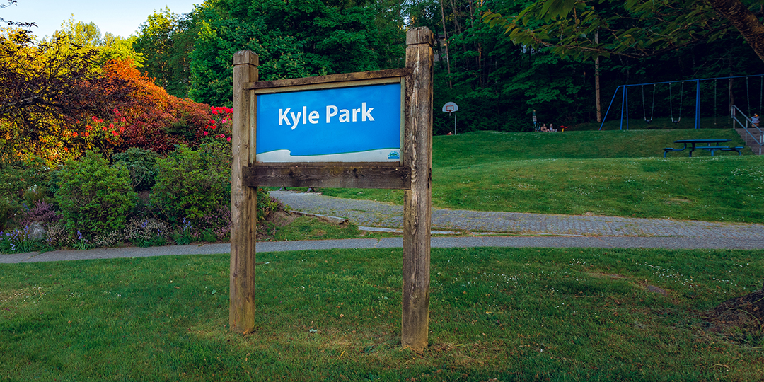 Green space with shrubbery in behind a blue sign that says Kyle Park