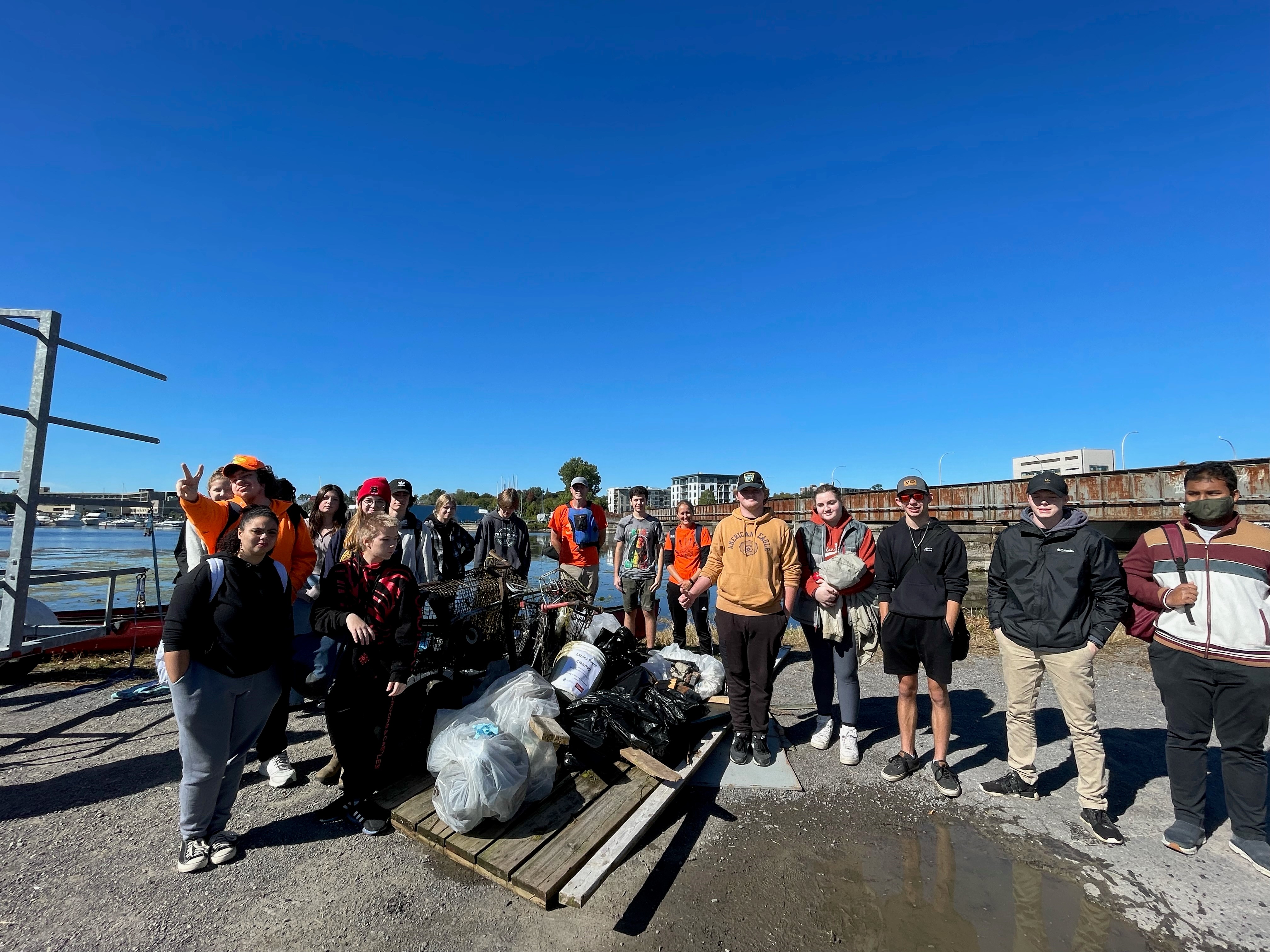 Group of volunteers participating in a clean-up event, standing proudly next to collected trash with a sunny blue sky and water in the background.