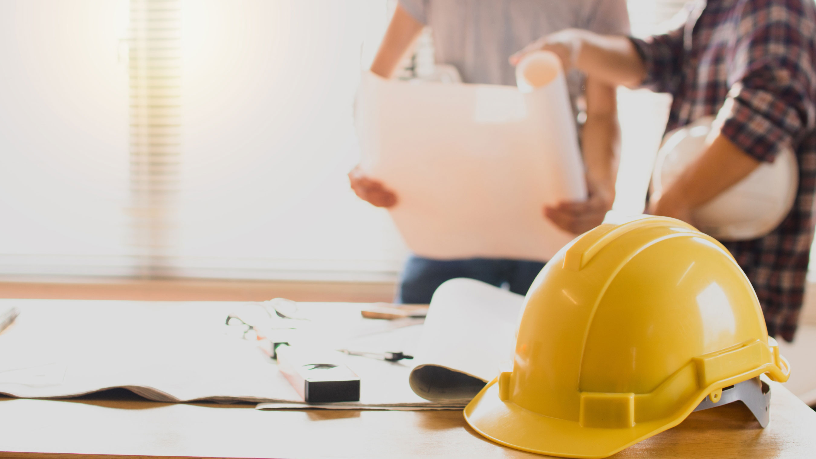 Two individuals examining blueprints at a work desk with various tools and a yellow hard hat visible in the foreground.