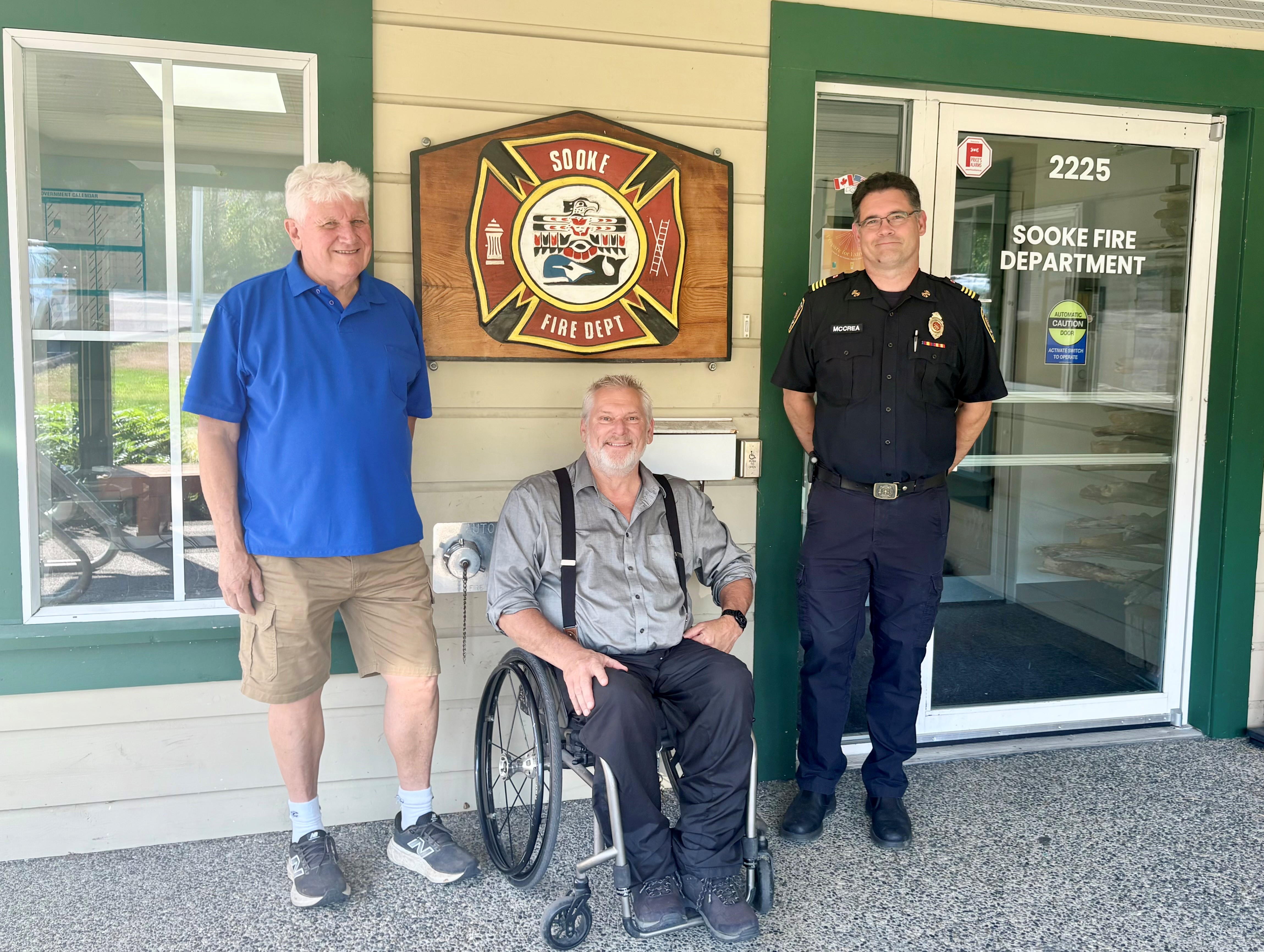 Deputy Mayor Al Beddows, MLA Dana Lajeunesse, seated in a wheelchair, and Deputy Fire Chief Chris McCrea pose outside the Sooke Fire Department.