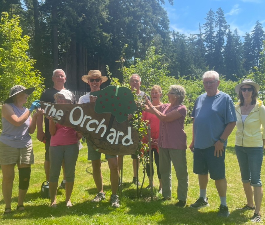 A group of people stand together outdoors in an orchard beside young fruit trees and a storage container during a Sooke Region Food CHI meeting on local food security and gleaning program expansion.