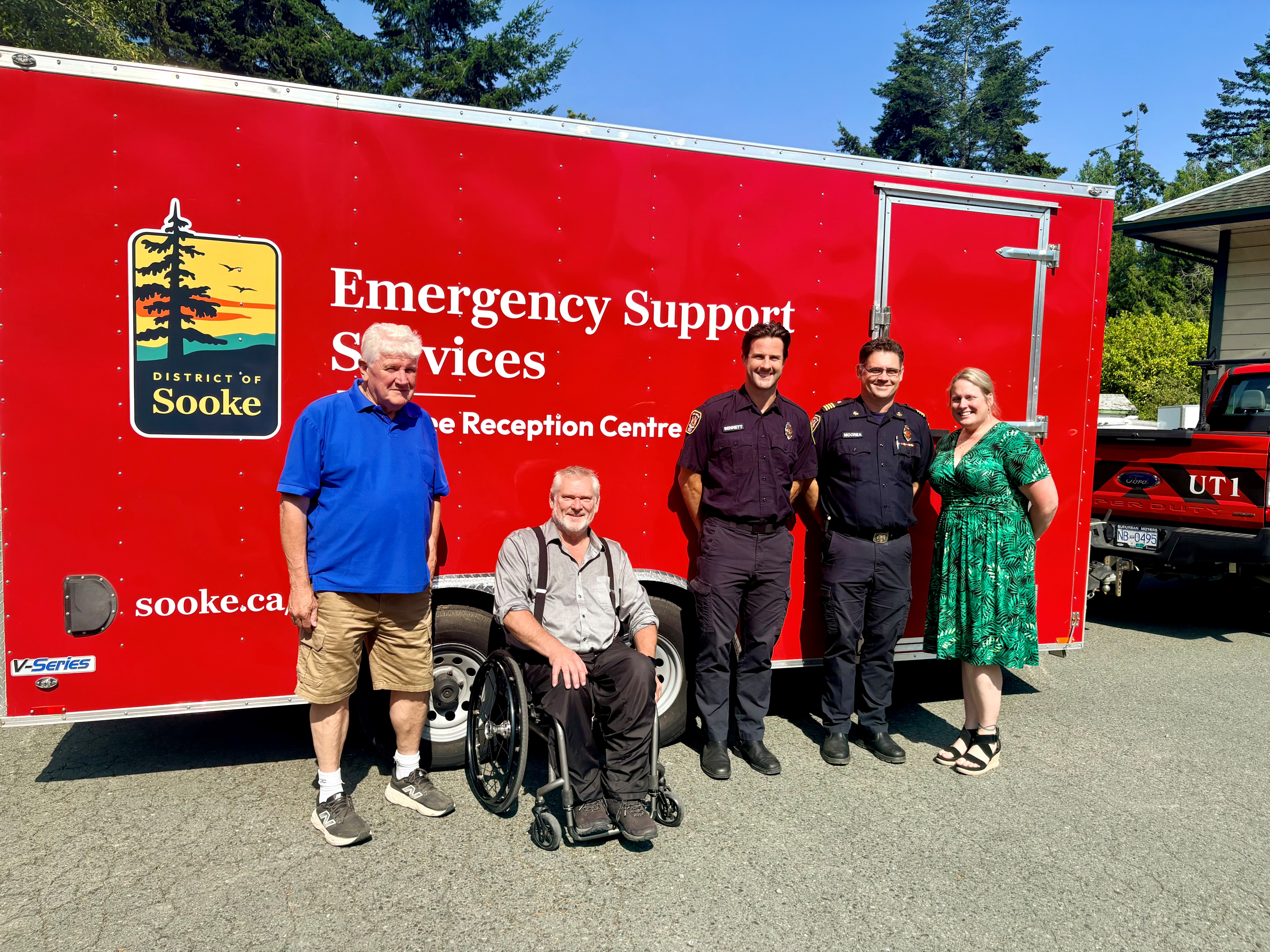 Five District and provincial representatives pose in front of the District of Sooke’s red Emergency Support Services Reception Centre Trailer: Deputy Mayor Al Beddows, MLA Dana Lajeunesse, Emergency Program Manager Ryland Bennett, Deputy Fire Chief Chris McCrea and CAO Raechel Gray.