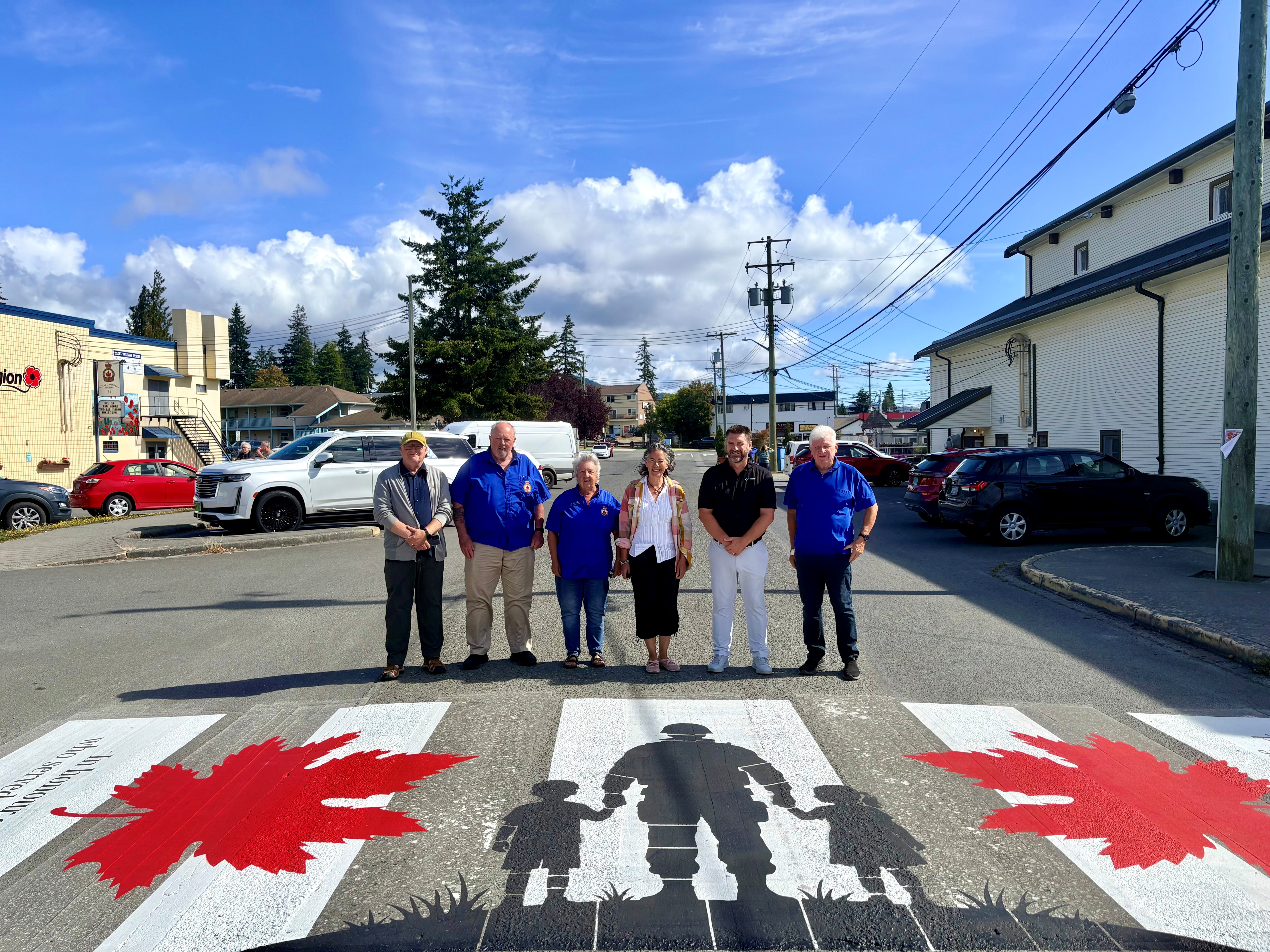Six representatives from the District of Sooke, Sooke Legion Branch 54 and Victoria Contracting stand behind the newly installed commemorative veterans crosswalk on Eustace Road, featuring red maple leaves and the silhouette of a service member holding the hands of two children.