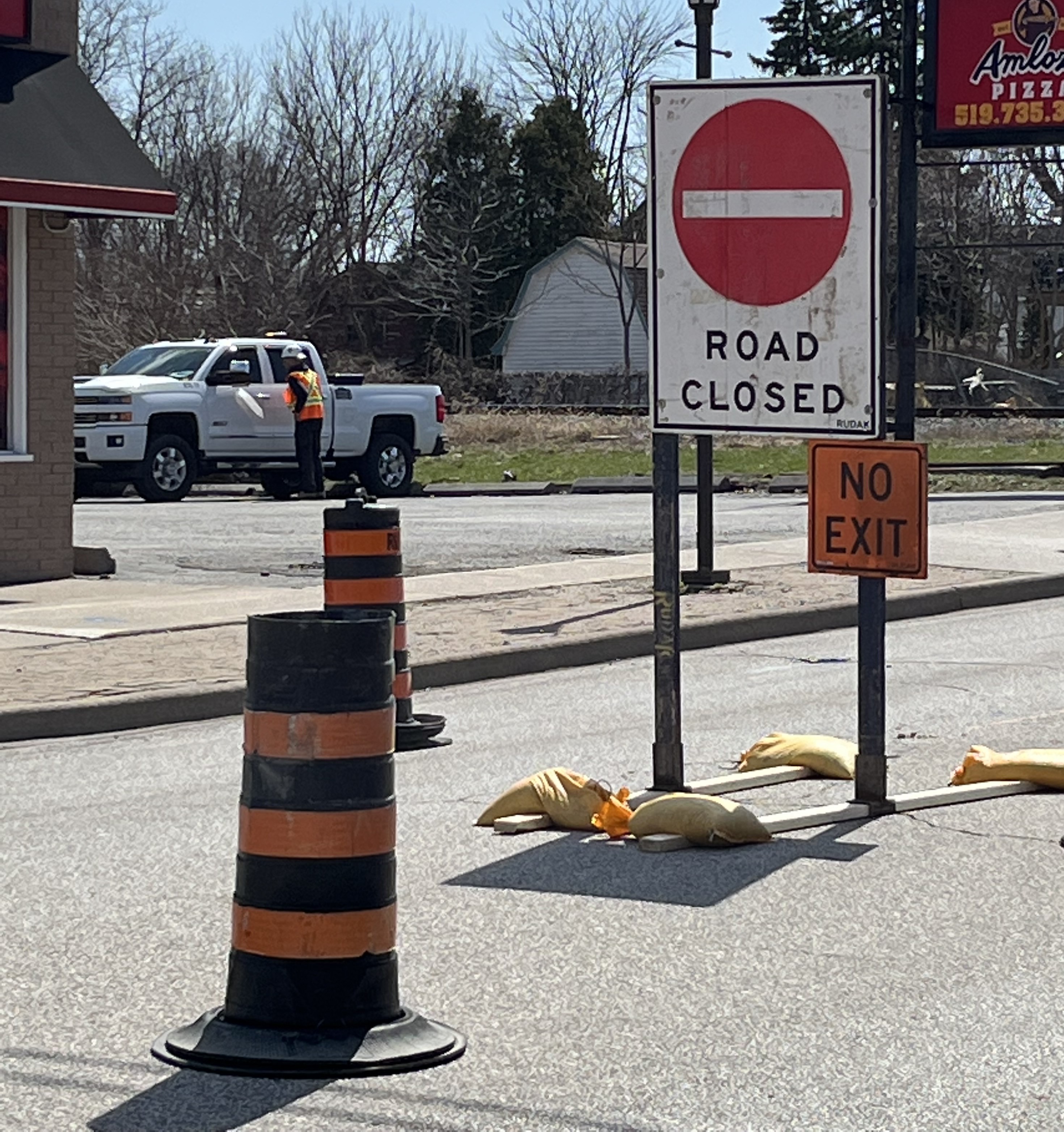 Two road closed sign displaying  "ROAD CLOSED" and "NO EXIT" on a street surrounded by trees, with an asphalt surface and visible vehicle traffic nearby.