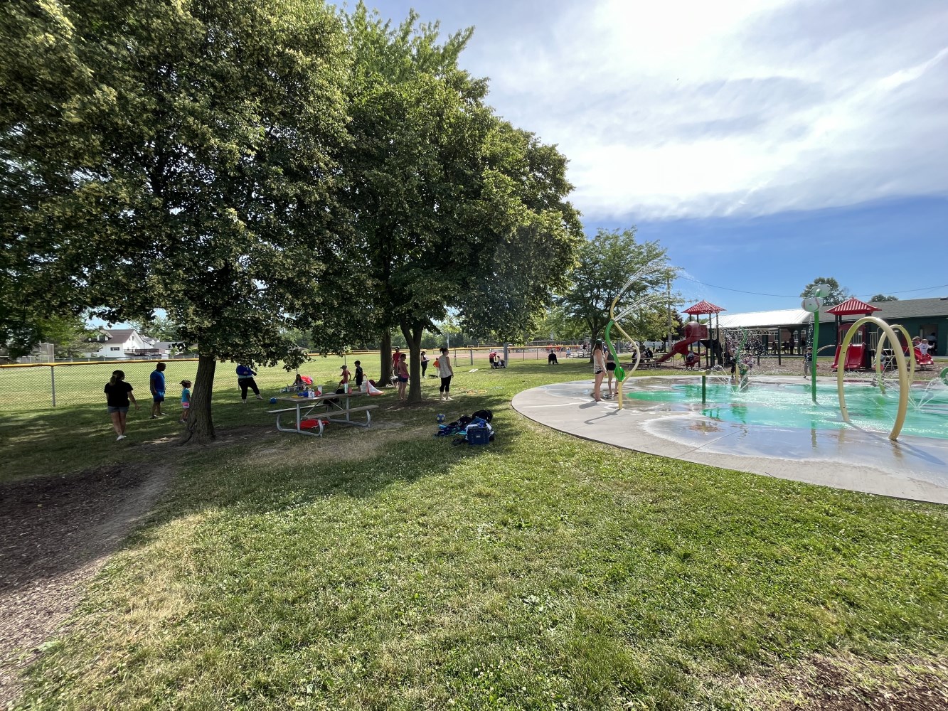 A diverse group of people enjoying a sunny day in a park, surrounded by lush green grass, trees, and fluffy clouds in the blue sky. There is a playground nearby and some water features visible in the background.