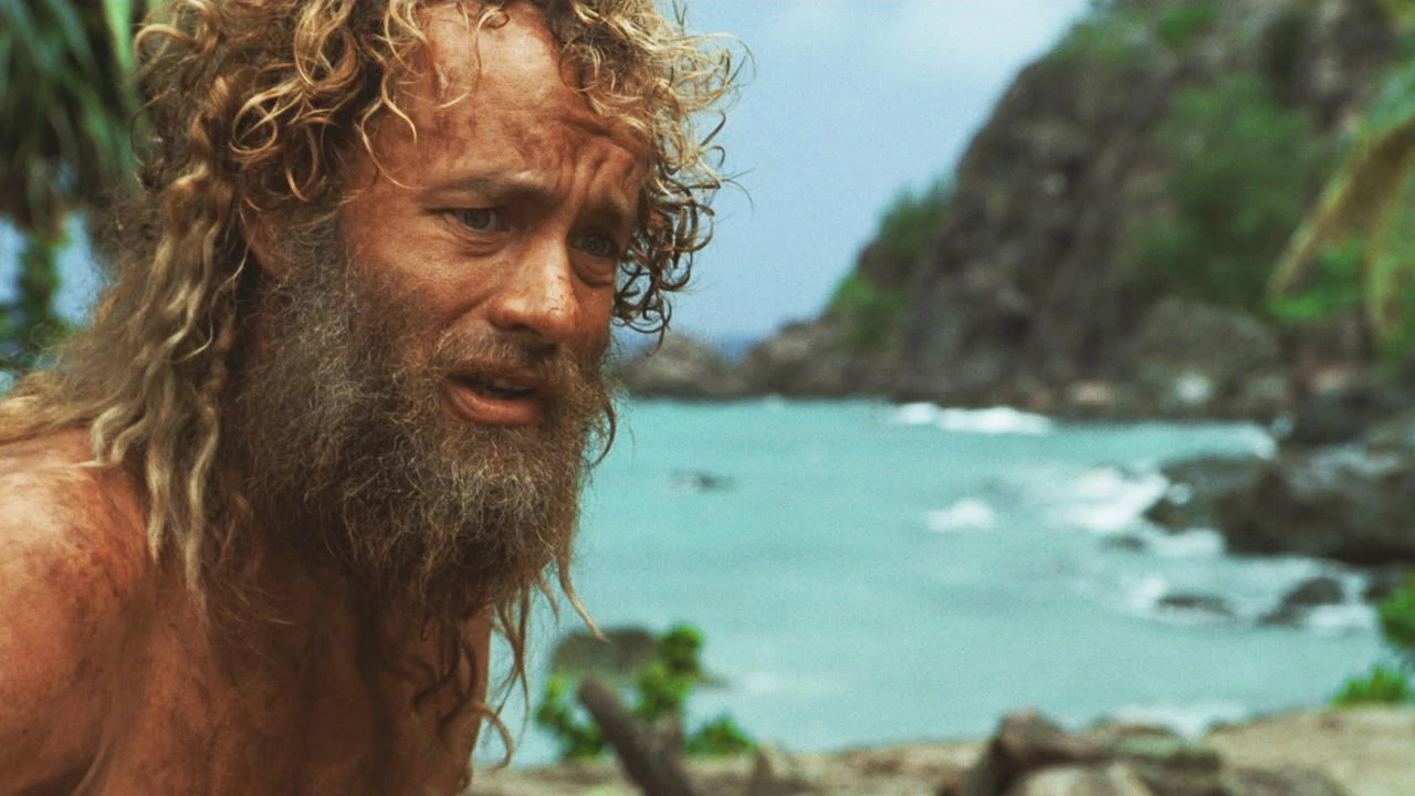 A person with long curly hair and a beard looks distressed on a beach, with rocky cliffs and the ocean in the background. 