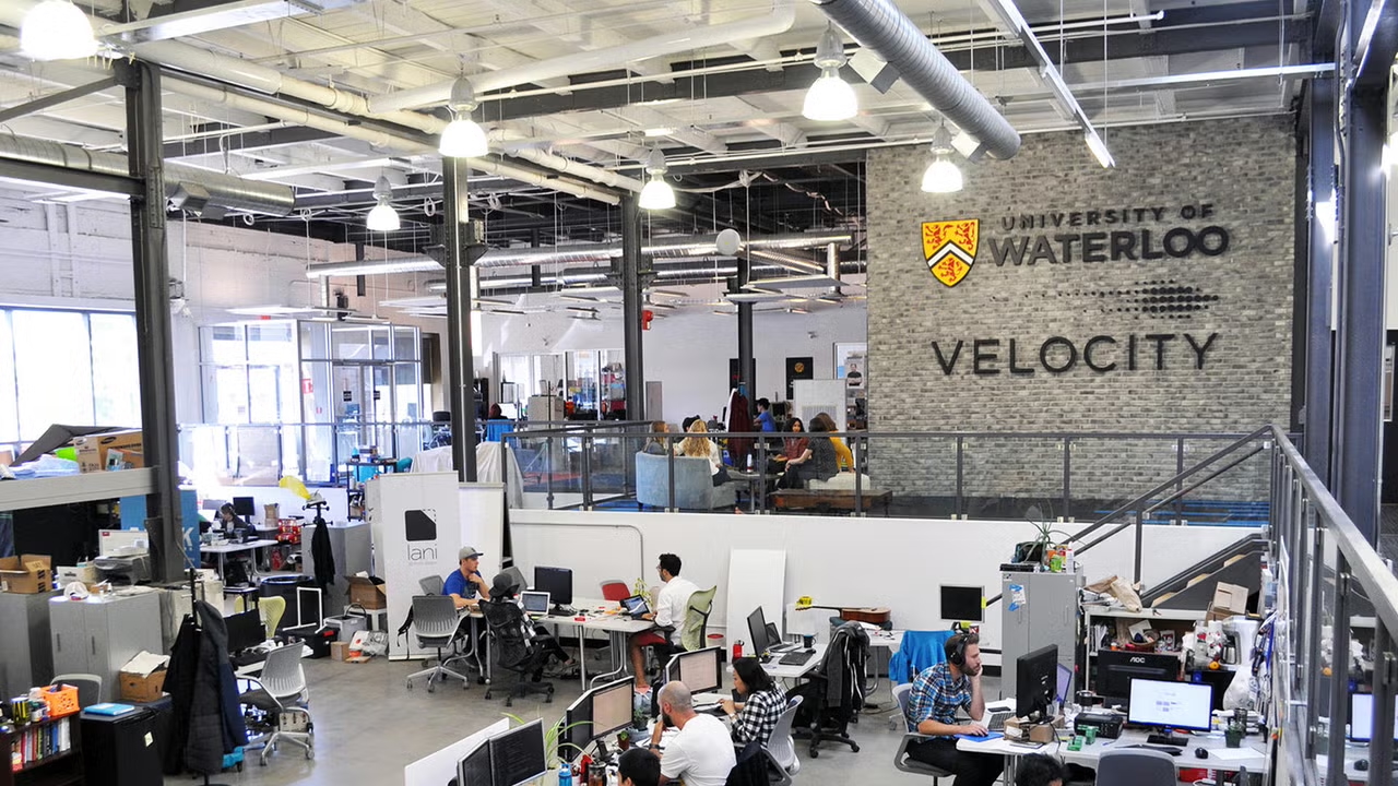 A modern open office space with people working at several desks and a sign on the wall with the University of Waterloo logo and text that says "Velocity".