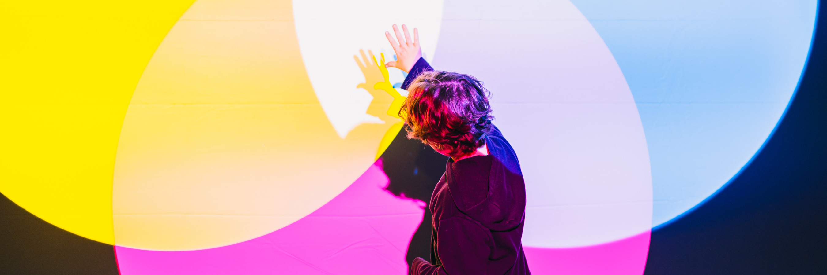 A person stands with one arm raised, interacting with overlapping colourful light projections against a dark wall.