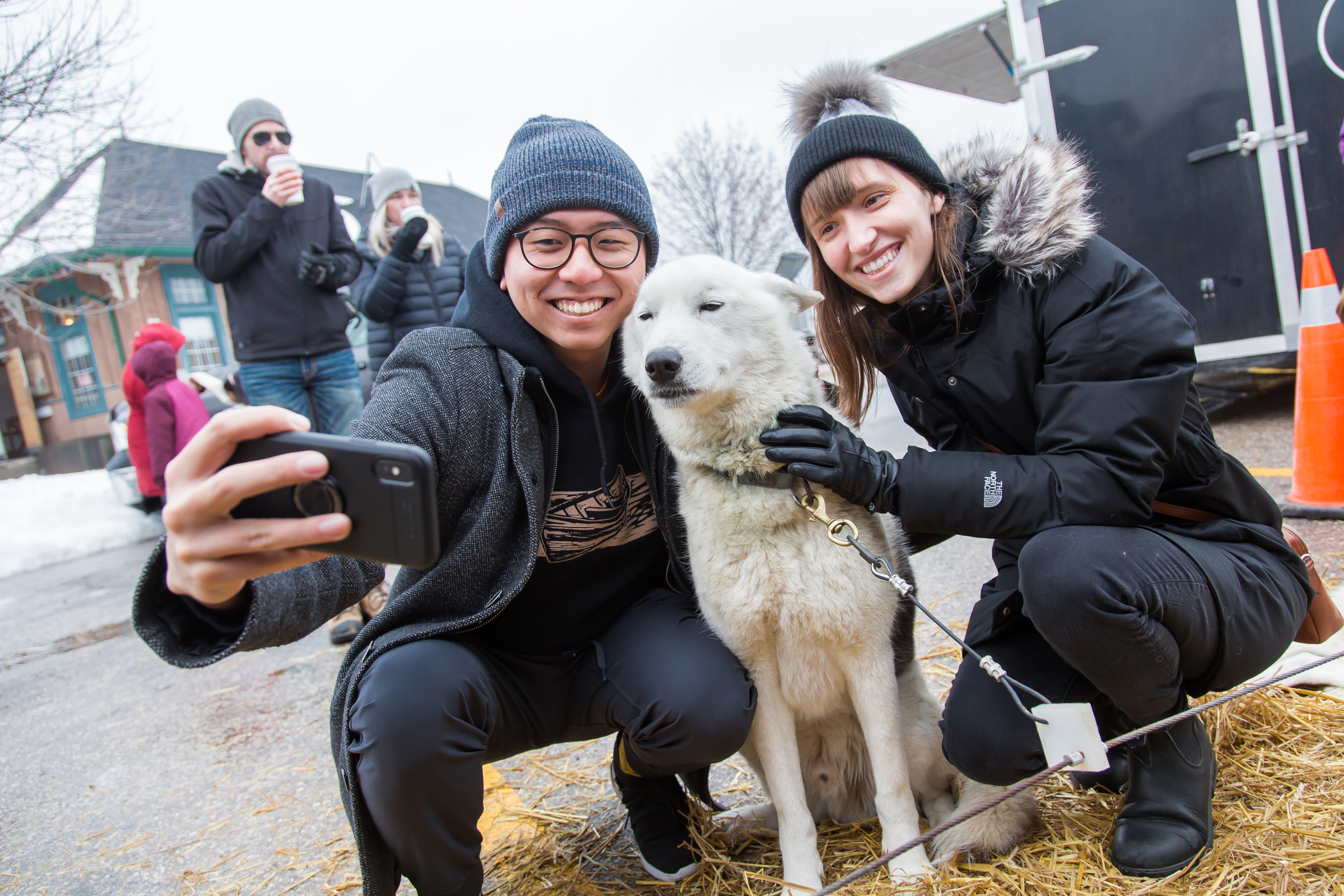 Two people wearing winter coats and hats, smile with a fluffy white dog at outdoor community event.