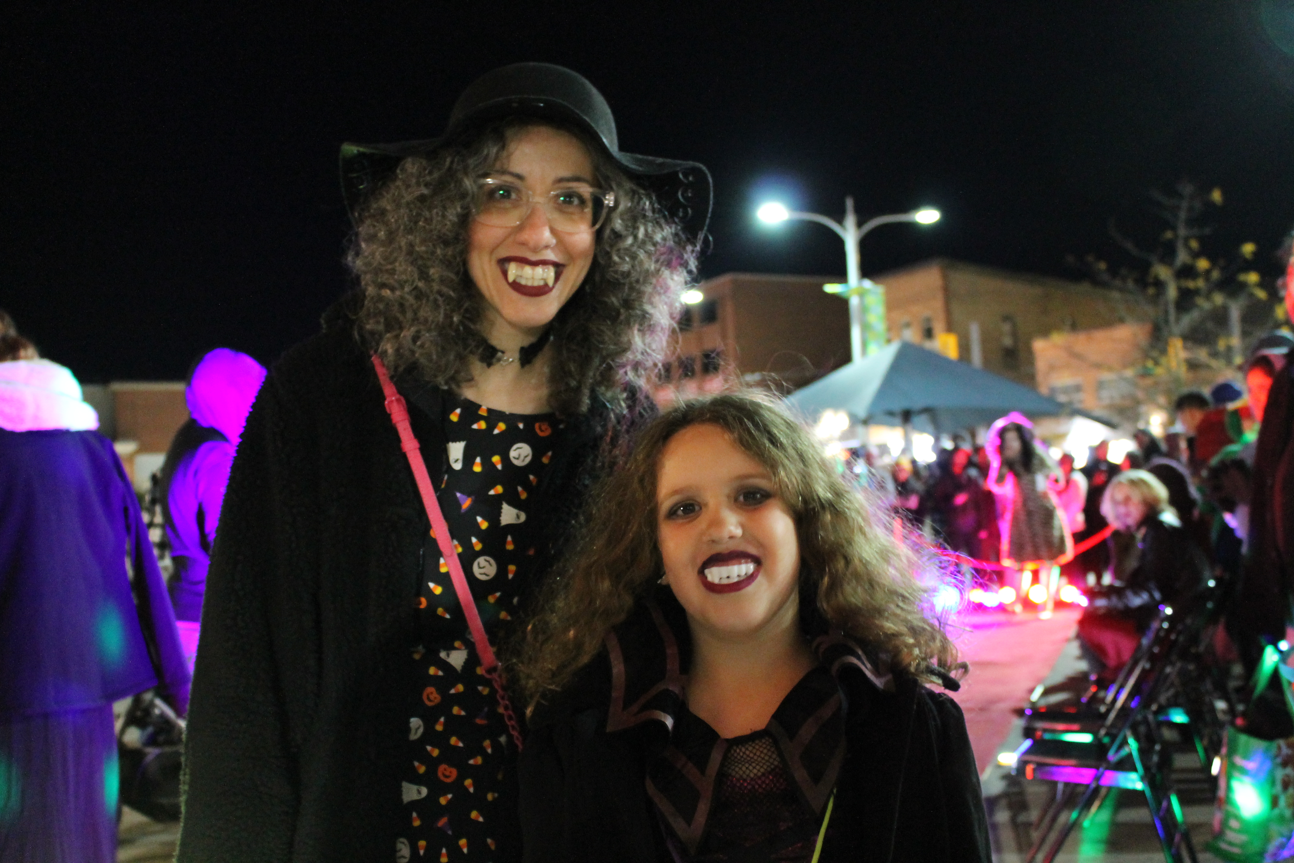 Two people smile at camera, wearing vampire teeth and clothes at evening event outdoors.