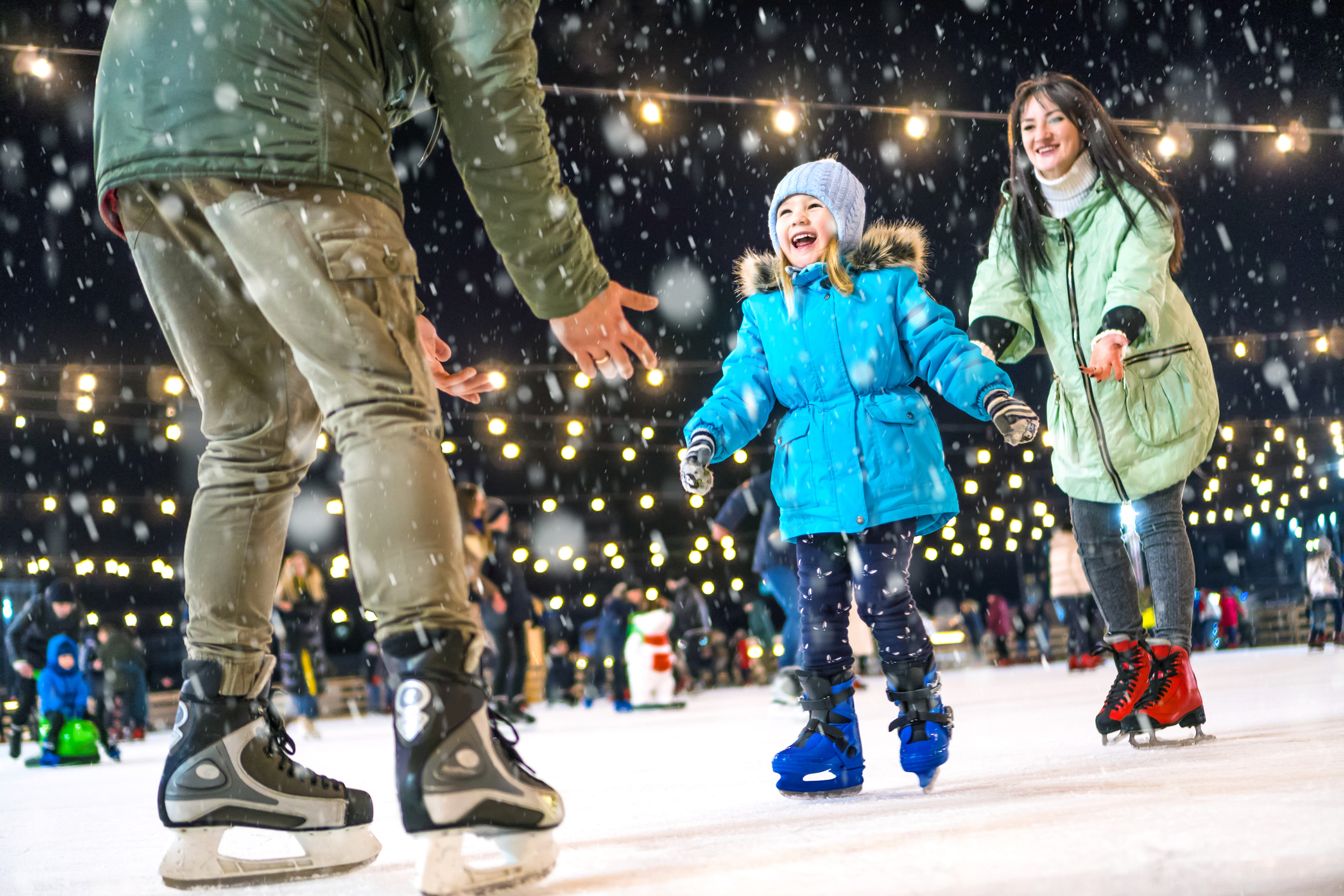 A child skates with two adults on an outdoor rink at night, smiling as snow falls.
