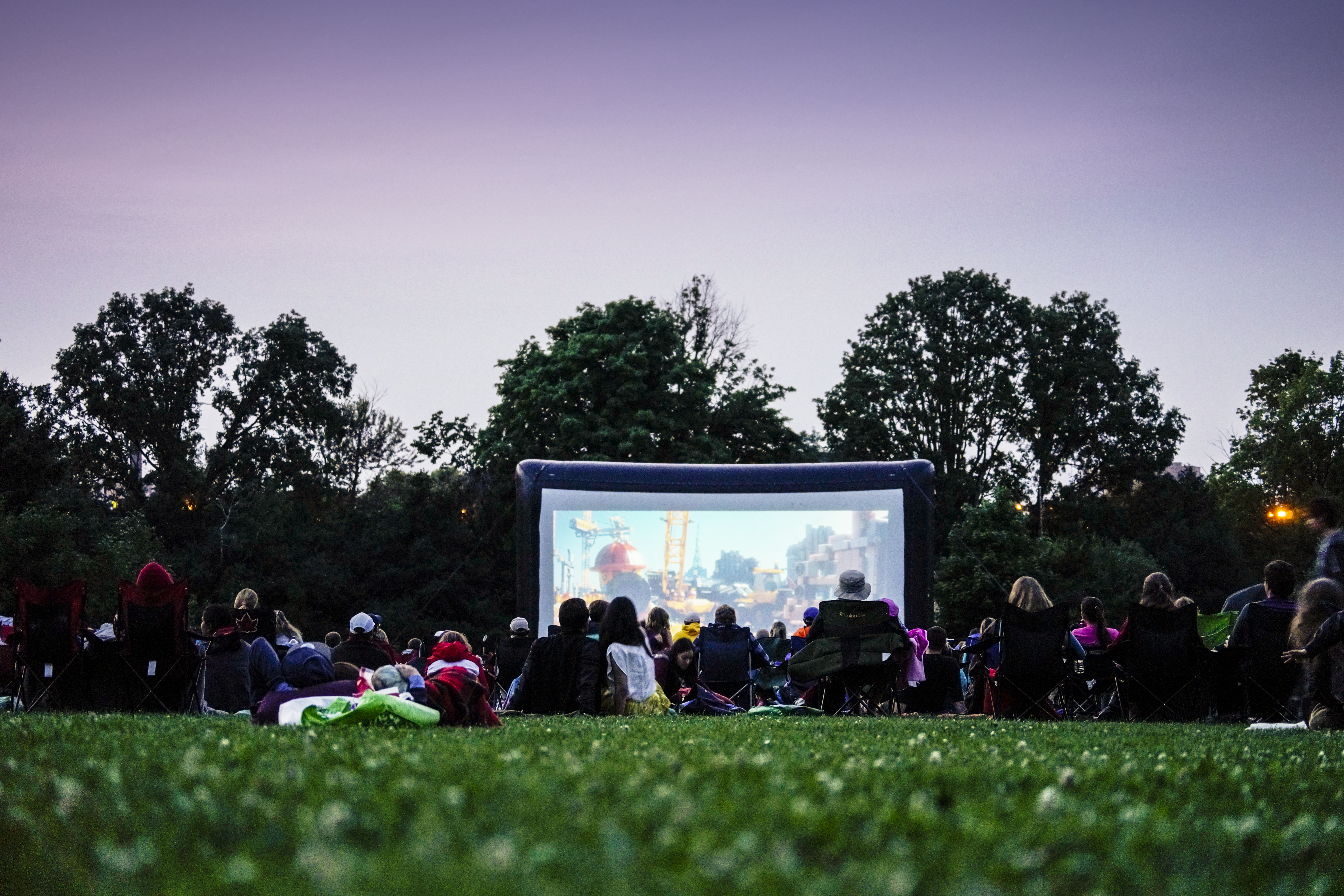 A crowd of people sitting on grass and chairs to watch an outdoor movie screening at dusk on a large inflatable screen, surrounded by trees.