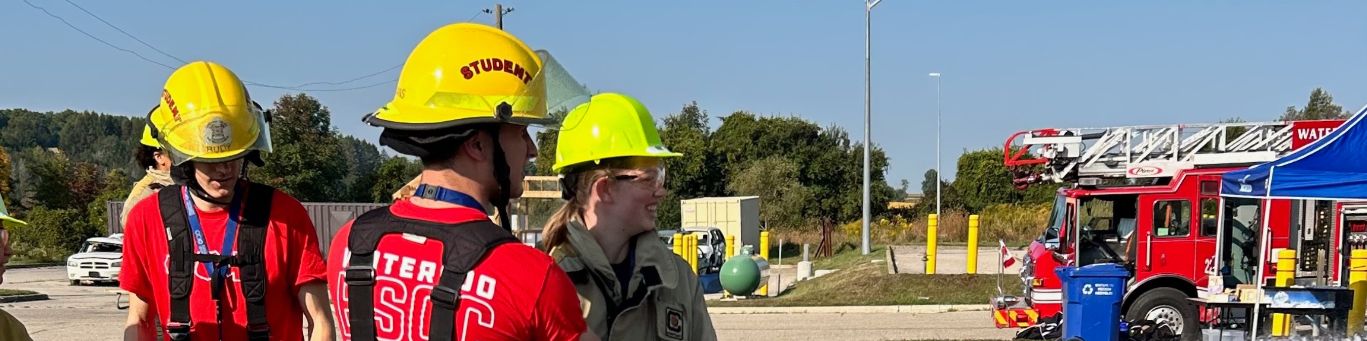 Group of people wearing yellow hard hats and safety gear stand together outdoors, speaking with a firefighter beside a red fire truck at a training or demonstration site.