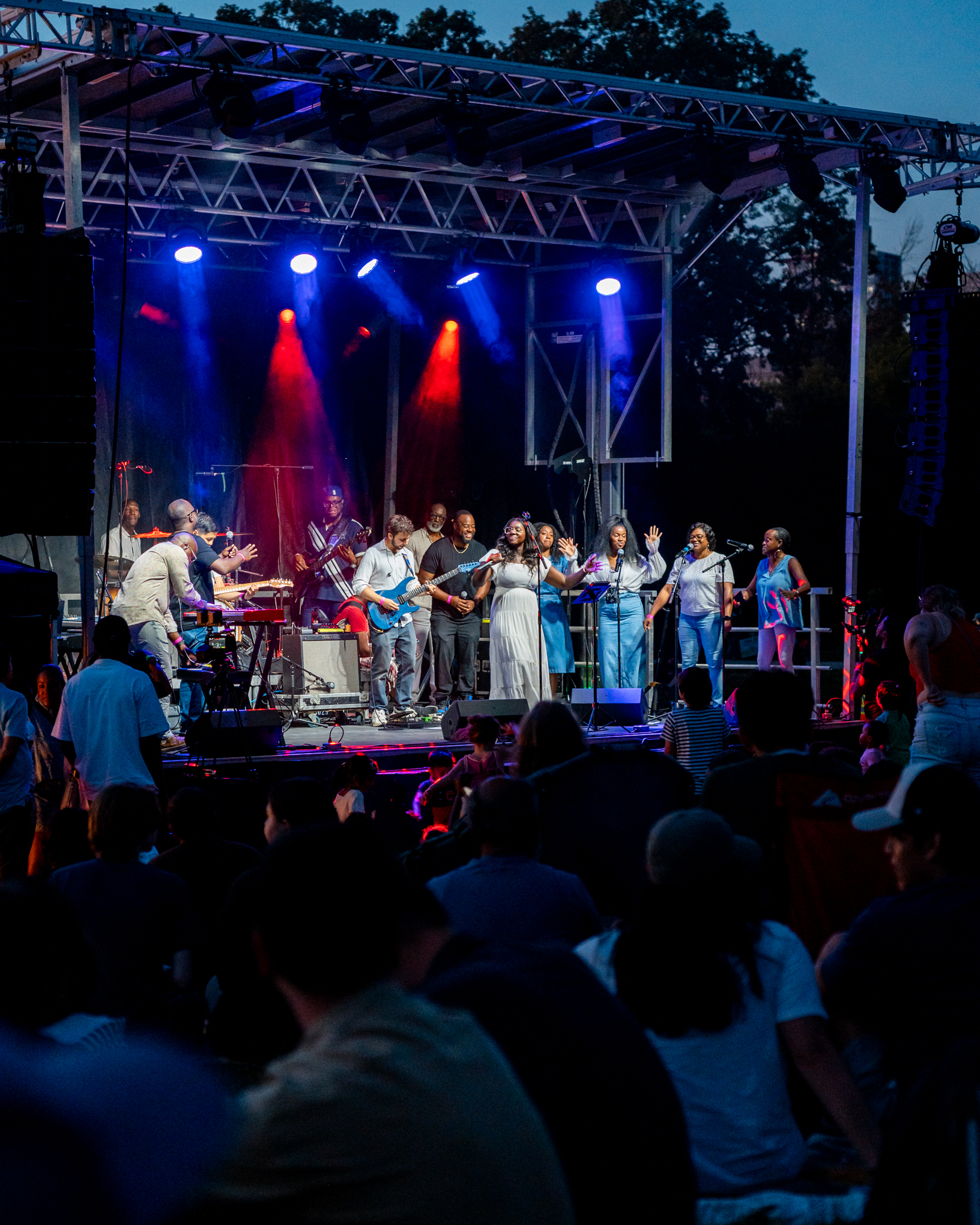 Musicians and singers perform under stage lights at an outdoor concert, with a crowd watching.
