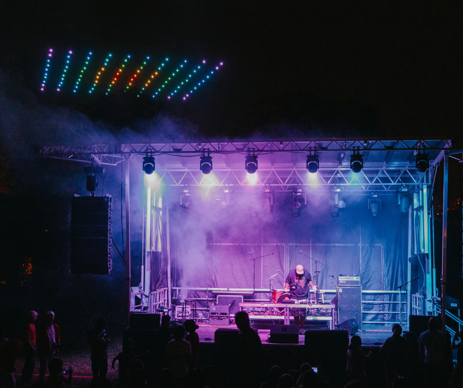 A night scene of an LED drone show above a large outdoor stage and crowd illuminated by lights.