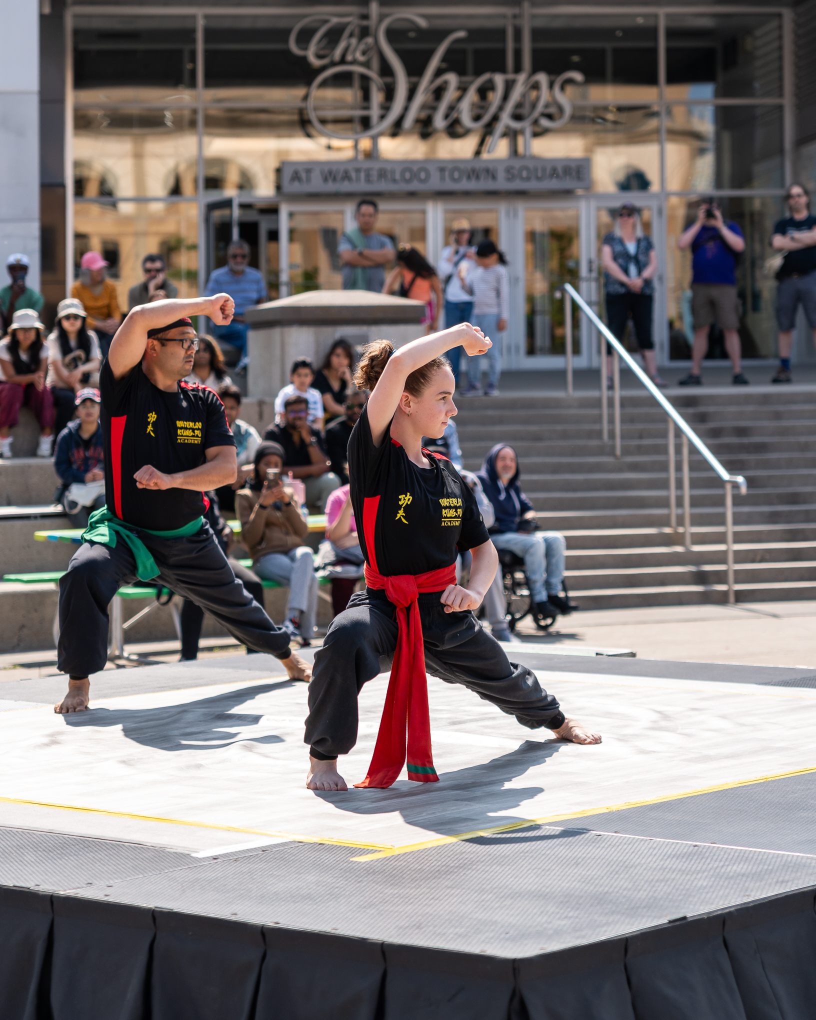 Two martial artists perform synchronized moves on an outdoor stage during a public event, dressed in matching uniforms with an audience seated watching nearby. 