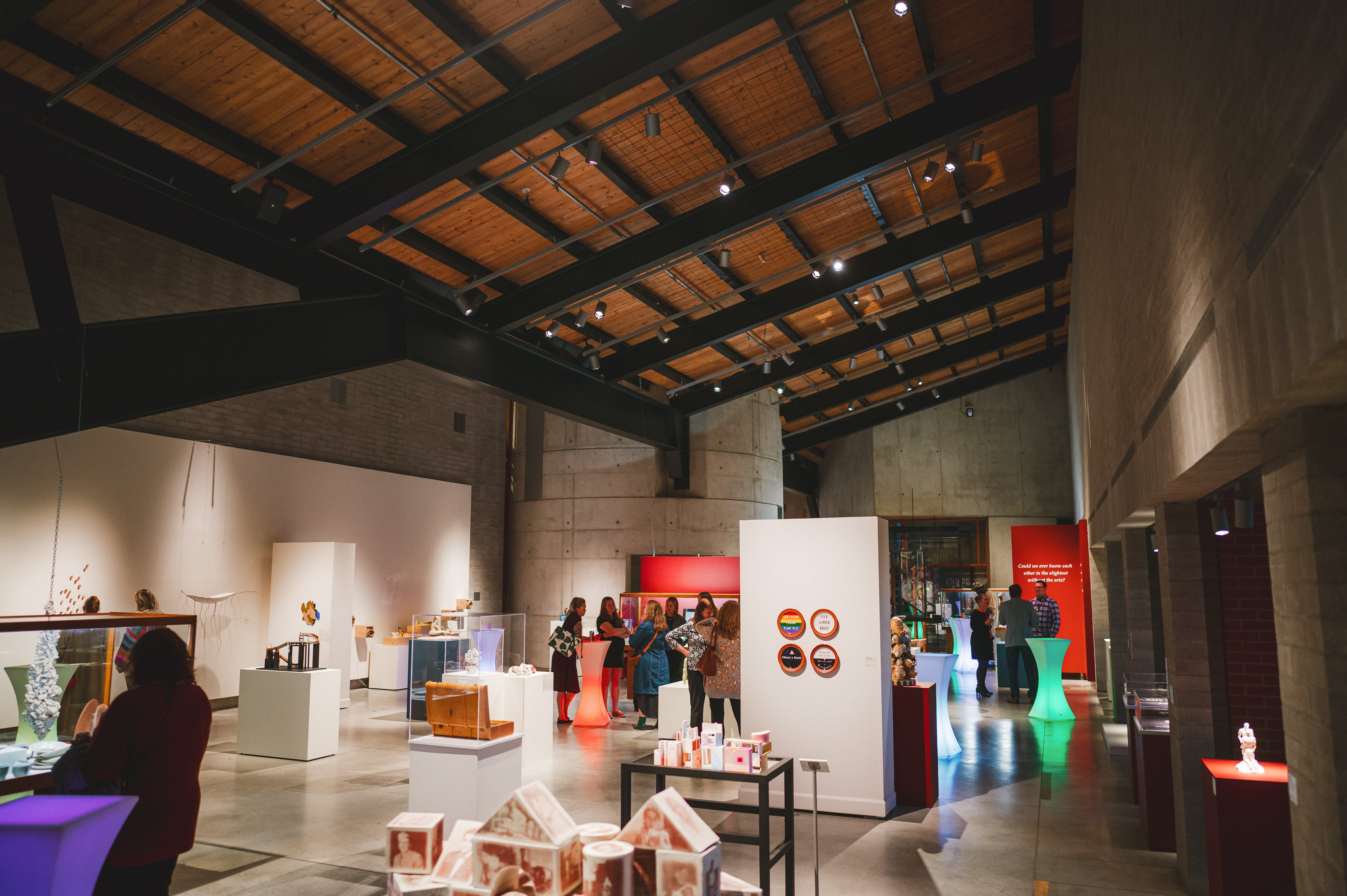 A modern art gallery interior with high ceilings and concrete walls featuring various artworks displayed on white pedestals and walls with visitors walking around.