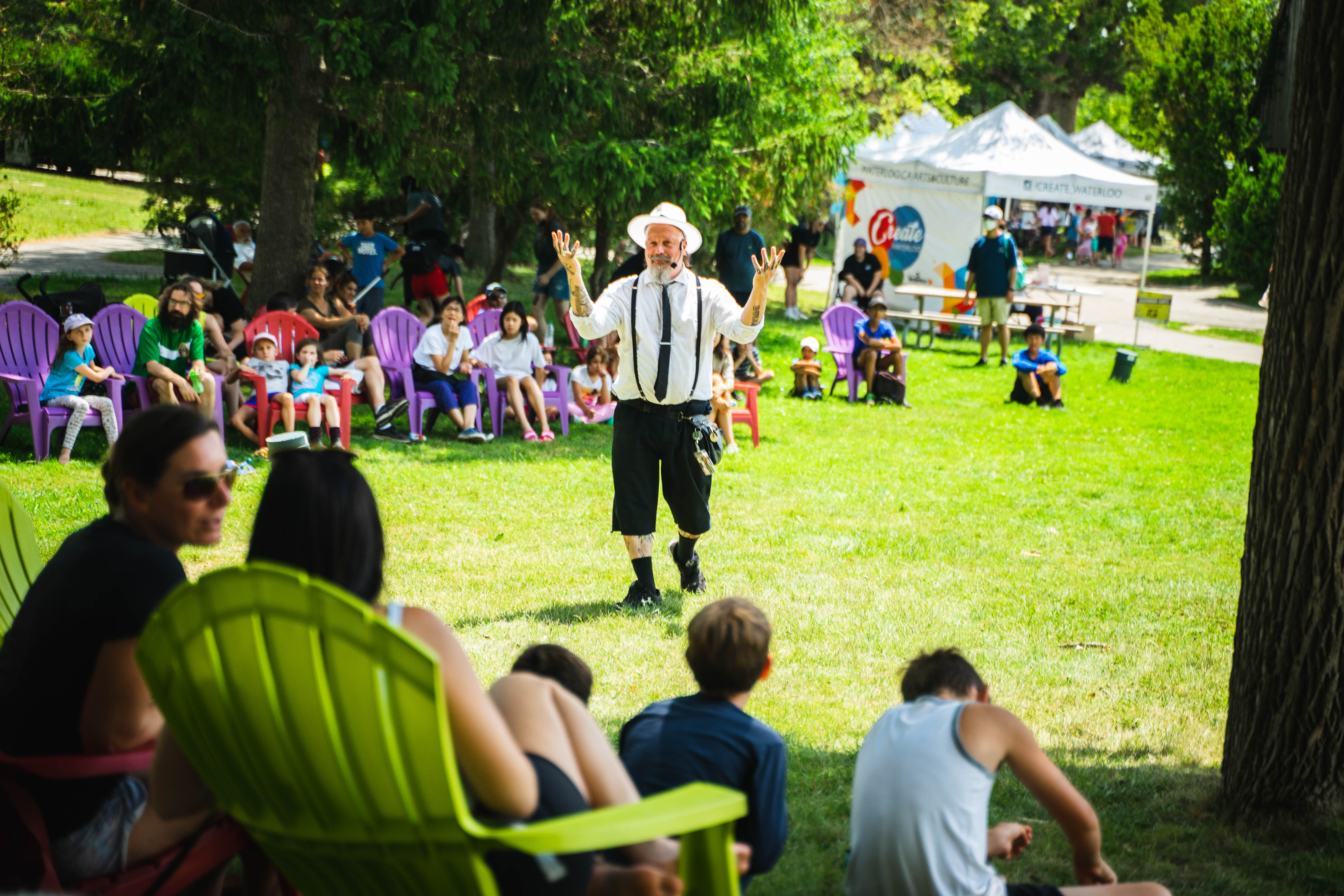 A lively outdoor event in Waterloo Park with people seated on colorful chairs, watching a performer in a hat and suspenders.