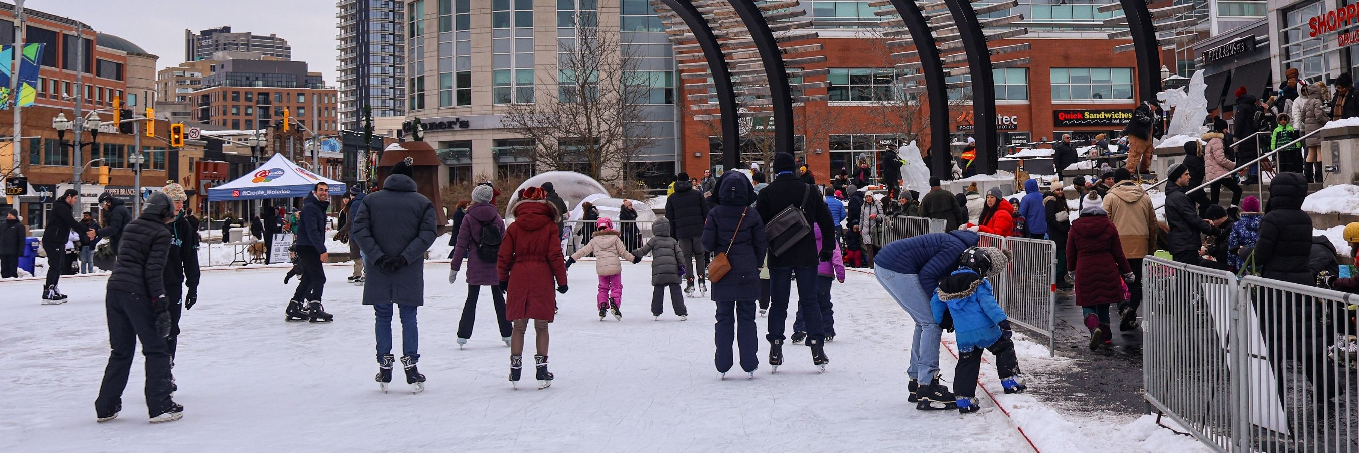 People of all ages skate on the outdoor rink at Waterloo Public Square on a winter day, with buildings and square features in the background.