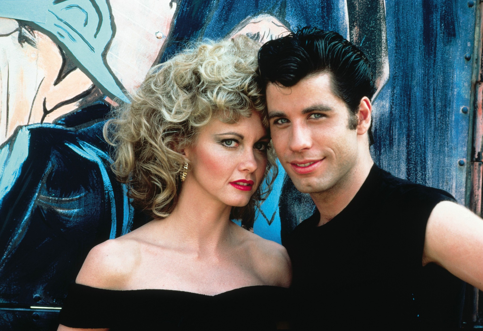 Two individuals are posing closely in front of a painted mural smiling slightly. 