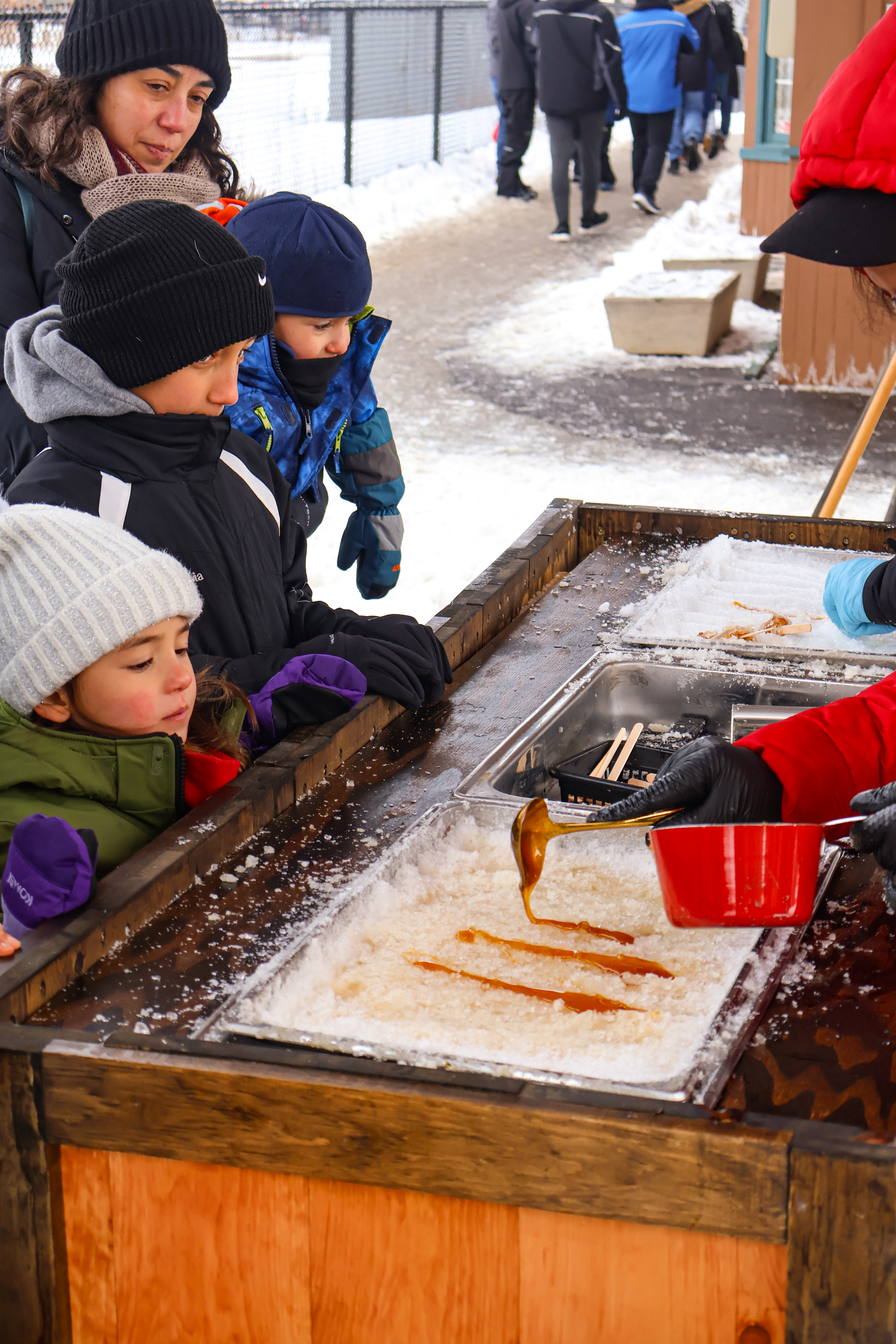 People gather around a wooden table as they watch a person pour maple taffy onto metal buckets of ice.