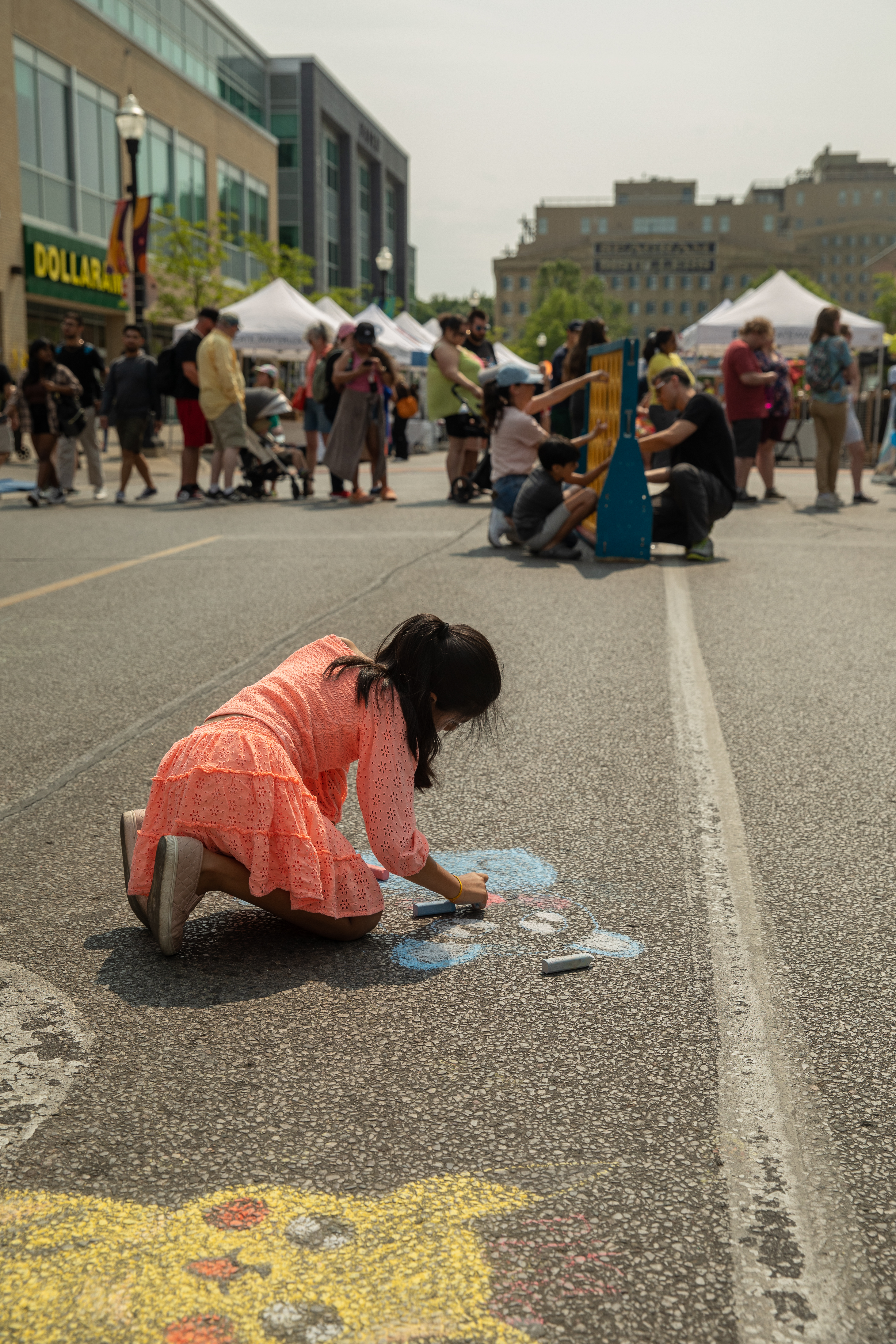 Child draws with chalk at a bustling street event with people playing various outdoor games in the background.