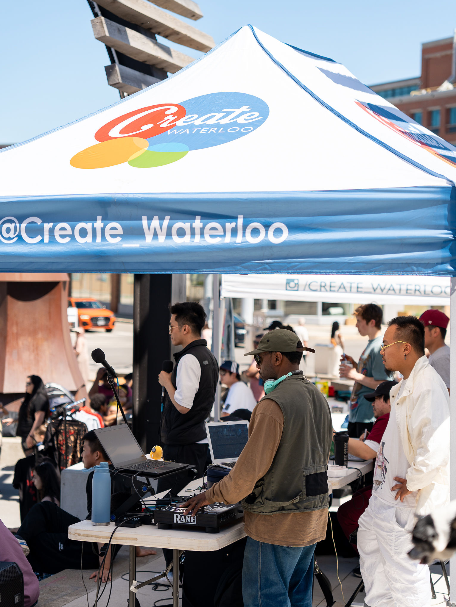Group of people using various music equipment and microphones stand under a tent titled '@CreateWaterloo' on sunny day.