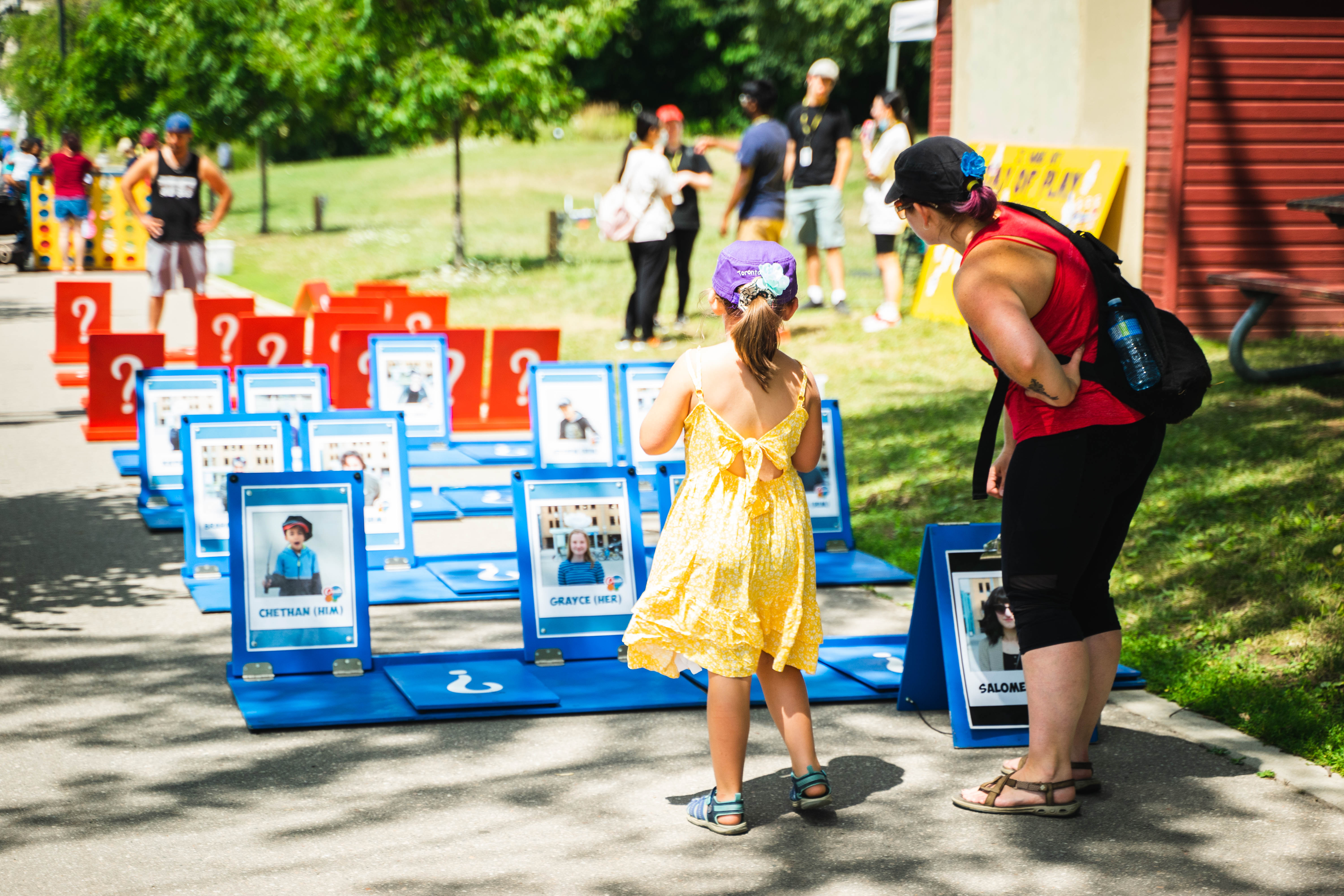 A person and a child are looking at a large outdoor game board of Guess Who on a sunny say in Waterloo Park with other people in the background.